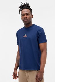 Man with dreadlocks wearing a navy blue t-shirt with a small cherry graphic and text, paired with beige pants, standing against a white background.