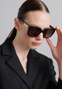 Černé oversized sluneční brýle s gradientovými skly, na což ukazuje zlaté logo "Moschino" na nožičkách. Model upravuje brýle, zatímco má na sobě černou sako.
