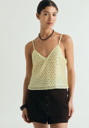 Mujer con una camisola color amarillo claro decorada con cuentas y tirantes finos, y una minifalda negra con botones, complementada con una pulsera y un collar negros.