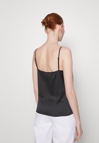 Camisola in satin nera con spalline sottili e regolabili, caratterizzata da una texture liscia e un orlo arrotondato, indossata con pantaloni bianchi.