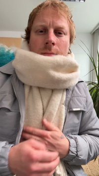 Beige wollen sjaal om de nek gewikkeld, met een zachte textuur. Grijze jas met ritssluiting gedeeltelijk zichtbaar. Binnenomgeving met planten.