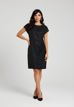 Mujer con cabello largo y oscuro, lleva pendientes de aro, vestido negro de manga corta con patrón texturizado y tacones negros, de pie con una mano en la cadera.