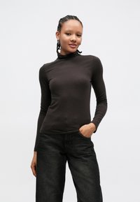 Donkerbruine, nauwsluitende coltrui met lange mouwen. Het model combineert het met losse zwarte jeans met zakken en een casual pasvorm.