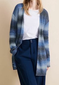 Stickad kofta i olika nyanser av blått, med öppen framsida, långa ärmar och ribbade ärmslut, bärs över en vit topp och marinblå byxor.