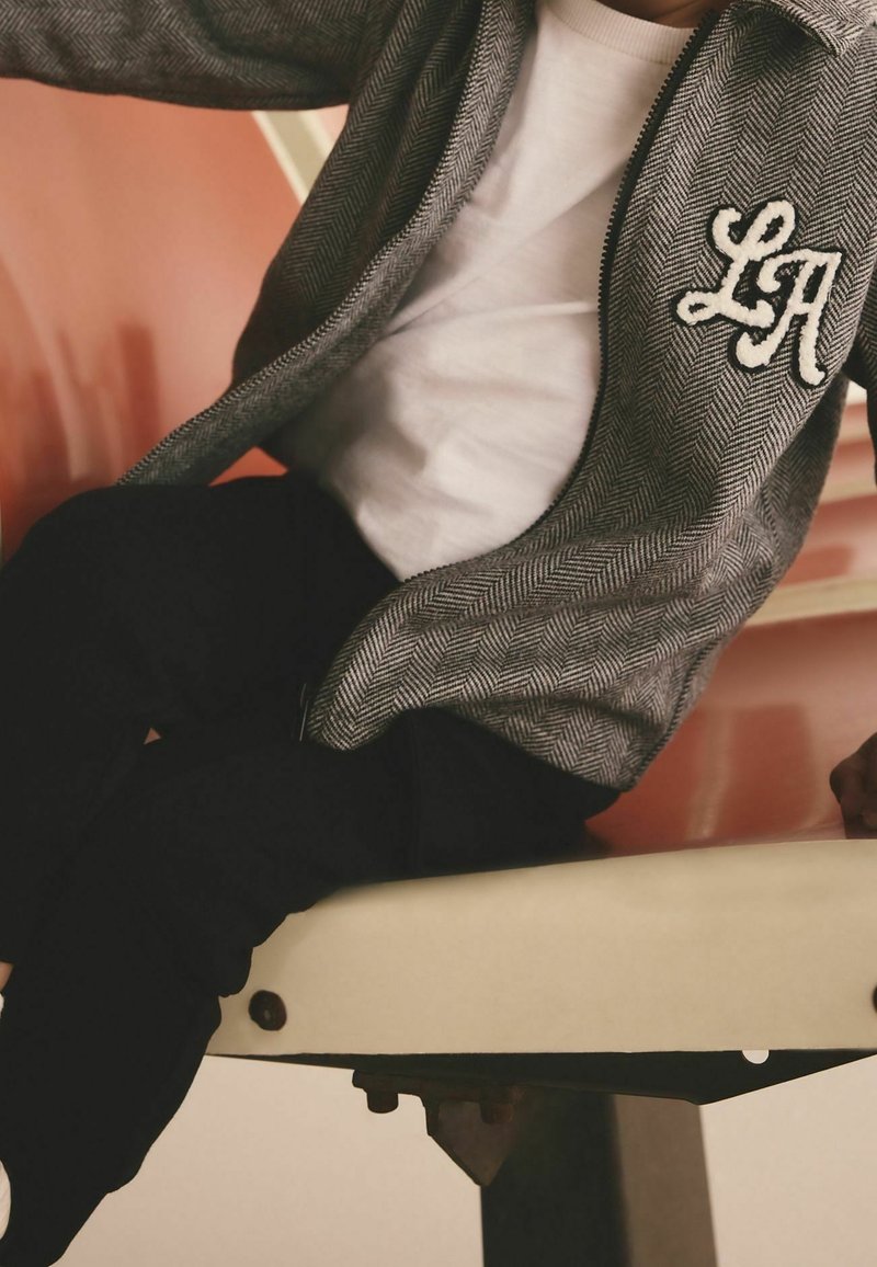 Enfant portant une veste grise à motifs avec un patch "LA", chemise blanche et pantalon noir assis sur un banc ou siège couleur crème.