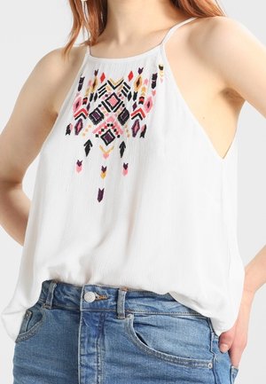 Weiße ärmellose Bluse mit mehrfarbiger geometrischer Stickerei auf der Brust, kombiniert mit hoch taillierten blauen Jeans aus Denim.