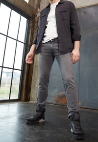 Homme portant un jean gris, des baskets montantes noires, un t-shirt blanc et une chemise noire déboutonnée, debout près d'une grande fenêtre dans un cadre industriel.