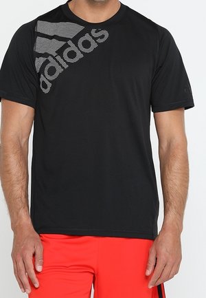 Mand iført en sort Adidas t-shirt med logo på skulderen og lyserøde røde shorts foran en ensfarvet baggrund.