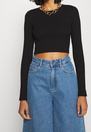Czarny prążkowany crop top z długim rękawem, połączony z wysokim stanem niebieskimi dżinsami w szerokie nogawki. Wyrazisty naszyjnik z ozdobnym złotym łańcuchem.