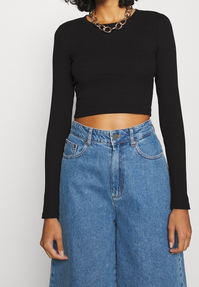 Mustansin riehumaton pitkähihaine crop-top, joka on yhdistetty korkeavyötäröisiin sinisiin farkkuleveyksiin. Huomionarvoinen kultaketju-aksento kaulakoru.