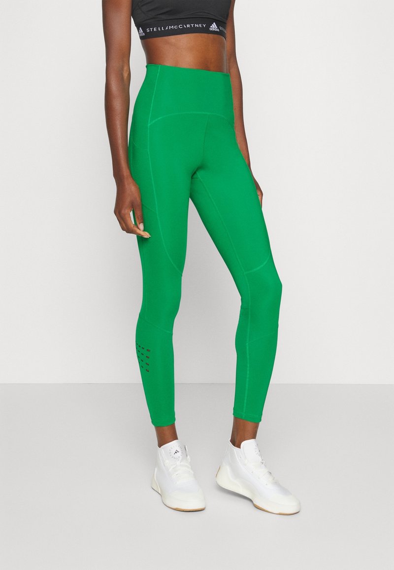 Kvinna med högmidjade gröna tights, svart sportbh med vit logga och vita höga sneakers mot en enkel bakgrund.