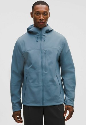 Uomo che indossa una giacca con cappuccio azzurro chiusa con zip, con tasche a zip sul petto e sui lati, in piedi contro uno sfondo chiaro e semplice.