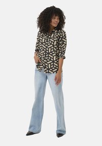 Schwarz-beige gemusterte Bluse mit geometrischem Design, Knopfdetails und hochgeschlagenen Ärmeln, kombiniert mit hellblauen Schlagjeans und schwarzen High Heels.