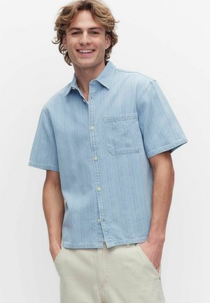 Jeune homme souriant, portant une chemise bleu clair à manches courtes avec poche devant et pantalon beige, mains dans les poches, fond blanc.