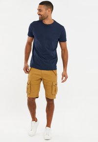 Navyblaues Baumwoll-T-Shirt kombiniert mit khakifarbenen Cargo-Shorts, die über zwei seitliche Taschen verfügen, und weißen Turnschuhen. Modell steht vor einem neutralen Hintergrund.