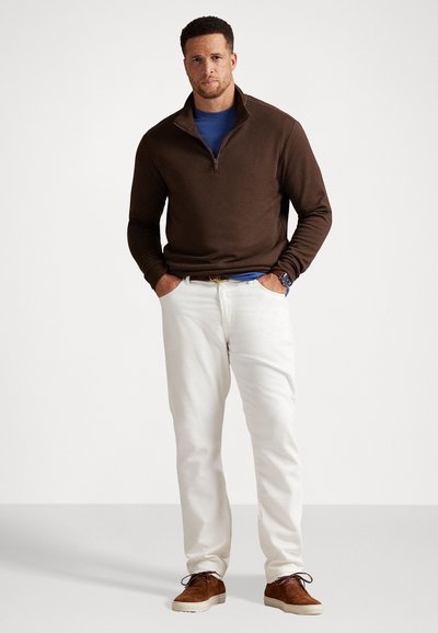Bruine ritstrui over een blauwe shirt, witte broek en bruine schoenen. Het shirt heeft een ronde hals, en de trui heeft een gladde textuur.