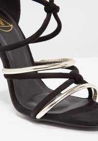 Sandale à talon haut en daim noir avec plusieurs brides entrelacées, ornée d'accents circulaires métalliques dorés et d'une semelle intérieure noire lisse.