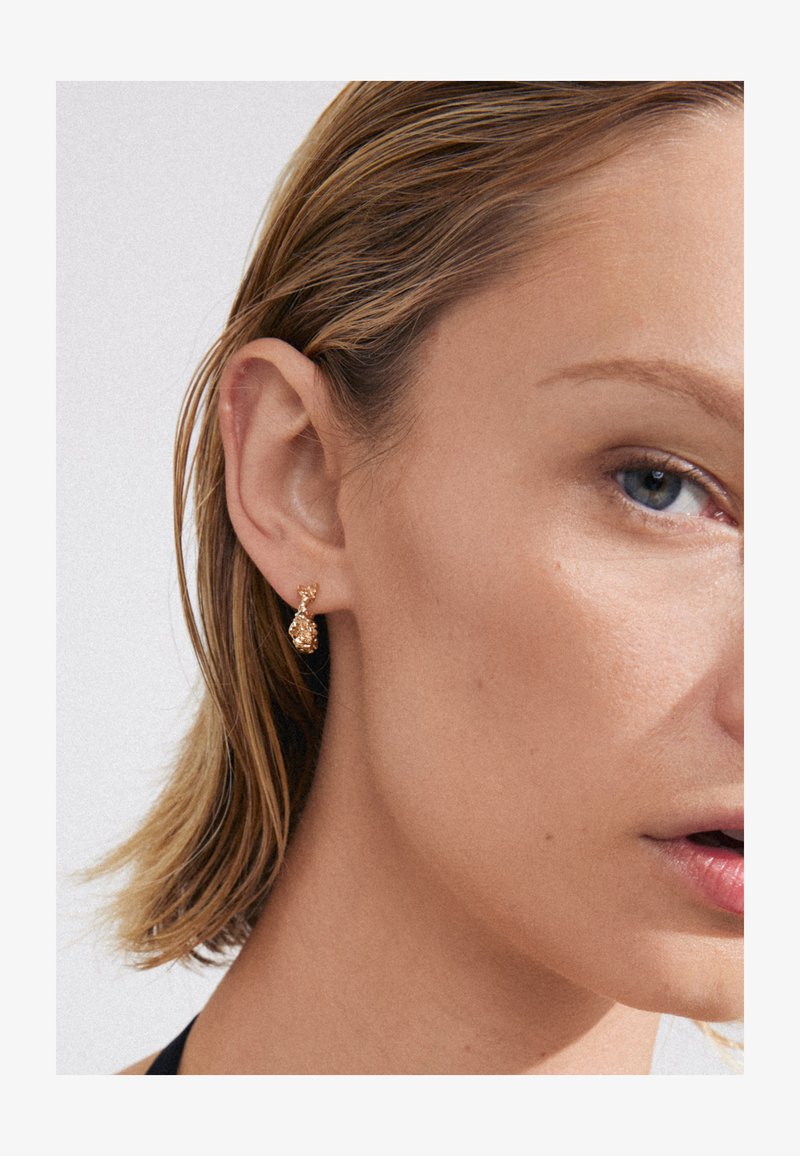 Boucles d'oreilles en or avec un design abstrait texturé. Les boucles d'oreilles sont fixées à un tige élégante et sont portées à une oreille.