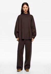 Blusa marrón texturizada de gran tamaño con alto cuello y mangas sueltas, combinada con pantalones de pierna ancha a juego y zapatos estampados.