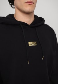 Black hoodie made of soft fabric, featuring a gold metal logo plate on the chest, with black drawstrings and gold metal tips.