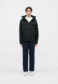 Ungdom som står iført en svart hettejakke med frontlomme, marineblå bukser, hvite joggesko og en hvit skjorte.