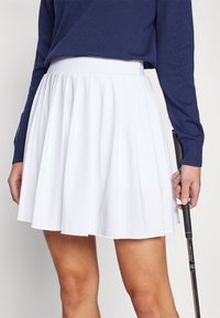 Bílá plisovaná sukně s hladkým páskem a rozšířeným siluetou, spárovaná s tmavě modrým svetr, držena golfovou holí.