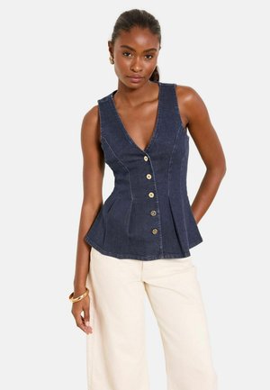 Naine kannab varrukateta tumesinist nööpidega teksatoppi ja kahvatult valgeid laia säärega pükse, aksessuaaridena kuldseid kõrvarõngaid ja käevõru.