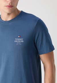 T-shirt in cotone blu con scollatura a girocollo e maniche corte. Presenta il logo ricamato bianco "TOMMY HILFIGER" e un piccolo dettaglio della bandiera rossa, bianca e blu.