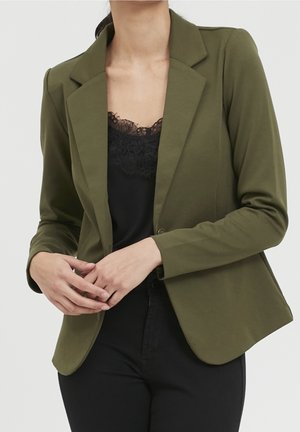 Vrouw draagt een olijfgroene blazer over een zwarte top met kanten rand, met handen gevouwen voor zich tegen een effen achtergrond.