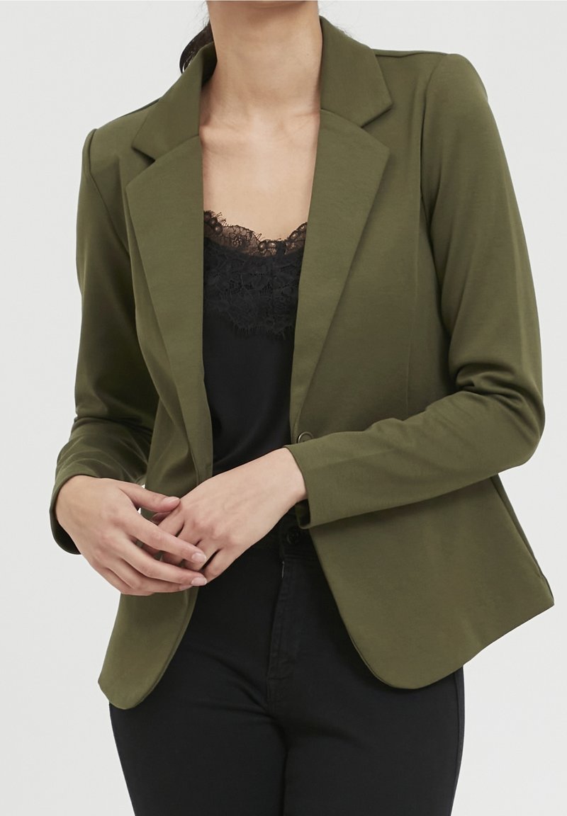 Femme portant un blazer vert olive sur un haut noir orné de dentelle, les mains jointes devant un fond uni.