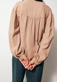 Henkilö, jolla on päällä pitkähihaista beigeä paitaa ja tummat housut, puristaa käsiään selän takana yksinkertaista taustaa vasten.