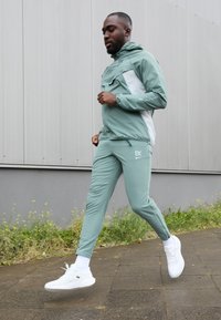 Mátově zelený sportovní teplákový komplet s kapucí, zipovou kapsou a kontrastními bílými detaily. Nošený s bílými teniskami a ponožkami do kotníků.