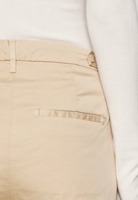 Pantaloni beige su misura realizzati in tessuto liscio. Presentano una tasca con zip e una chiusura con bottone. Accentuati da una leggera lucentezza.
