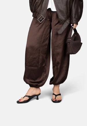 Femme portant un pantalon en satin marron clair ample avec des poignets élastiques, des sandales à talons noires, une veste en cuir marron foncé, et tenant un sac à main tissé assorti.