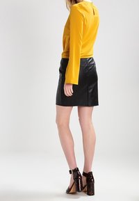 Žena v hořčičově žlutém dlouhém rukávu, černé kožené mini sukni a botách na blokovém podpatku s ankle-strap ve želvovinovém vzoru, stojící bokem.