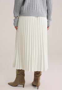 Persona, kas tērpusies pelēkā ribotā džemperī un baltā plīšveida midi svārkos, papildinot ar brūnām zamšādas zābakām ar vidēja augstuma papēdi, stāv uz gaiša fona.