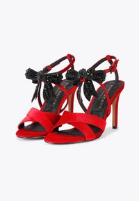 Czerwone satynowe sandały na wysokim obcasie z czarnymi koralikowymi kokardkami, otwarty przód, oraz smukły pasek na kostce. Teksturowana czarna wkładka.