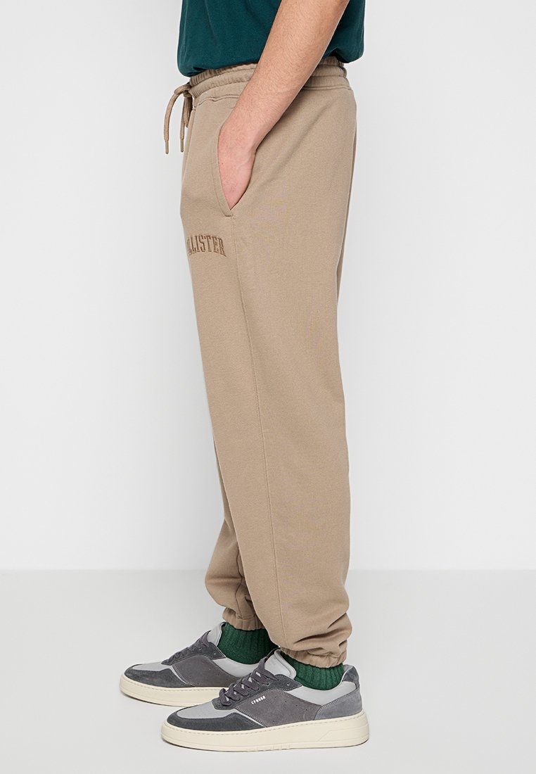 Beżowe spodnie dresowe z elastycznym pasem i sznurkiem, z bocznymi kieszeniami i haftowanym logo "Hollister" w kolorze brązowym.