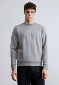 Γκρι φούτερ με υφασμάτινο, ανάγλυφο λογότυπο "BOSS" στο μπροστινό μέρος. Διαθέτει στρογγυλή λαιμόκοψη και ριμπ τελειώματα στα μανίκια και την άκρη. Απλός, λιτός σχεδιασμός.