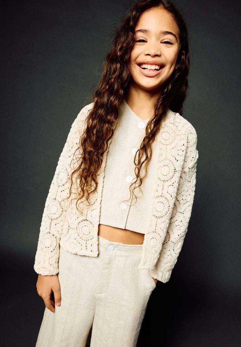 Jeune fille souriante aux longs cheveux bouclés, portant un cardigan en crochet crème, un haut court boutonné et un pantalon assorti, sur fond sombre.