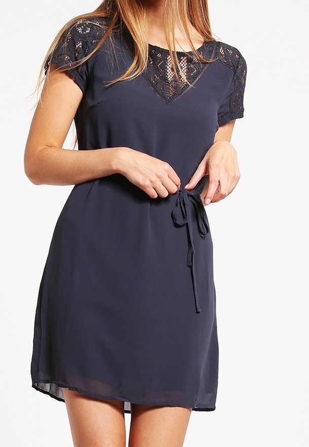 Žena upravuje pás na šaty s krátkým rukávem v námořnické modři s krajkovými detaily na výstřihu a rukávech.