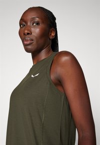 Confident woman with braided hair wears sleeveless olive green top with white logo, looking slightly upward against plain background.