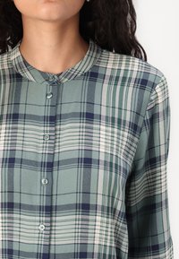 Grønn rutete skjorte med rund krage, trykknapper og en blanding av mørk marineblå, hvite og grønne striper i et strukturert stoff.