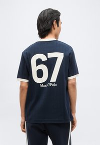 Άνδρας που φοράει μπλε κοντομάνικο πουκάμισο με λευκό «67» και το κείμενο «Marc O’Polo» στην πλάτη, στέκεται μπροστά σε απλό ανοιχτό φόντο.