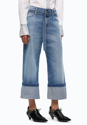 Mujer viste vaqueros azules anchos y cortos con grandes puños y un cinturón de tela a juego, combinados con tacones negros puntiagudos con cordones.
