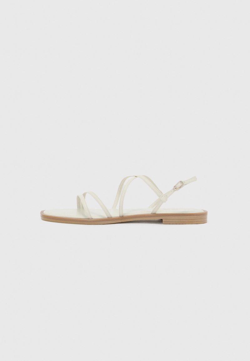 Sandale plate beige avec de fines lanières et une petite boucle sur la bride de cheville, présentée sur un fond clair uni.