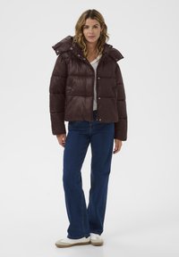 Brūns pūkains mētis ar kapuci, teksturēta auduma, kvadrāta formas, ar pogām aizdarāms un ar elastīgām aprocēm. Saskaņots ar ziliem džinsiem un baltiem apaviem.