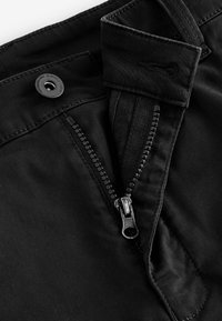 Los pantalones negros presentan un cierre con cremallera y un botón, con una cremallera metálica plateada, y una textura de tela mate y suave con detalles cosidos.