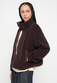 Bordeauxroter Fleece-Pullover mit hohem Kragen, Reißverschluss und Seitentaschen. Strukturierte Oberfläche und lockerer Sitz, getragen über einem hellen Oberteil.