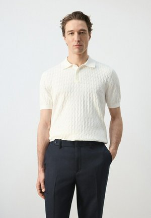 Mężczyzna w białej, krótko rękawowej, dzianinowej koszulce polo i ciemnogranatowych spodniach stoi z jedną ręką w kieszeni na jednolitym tle.